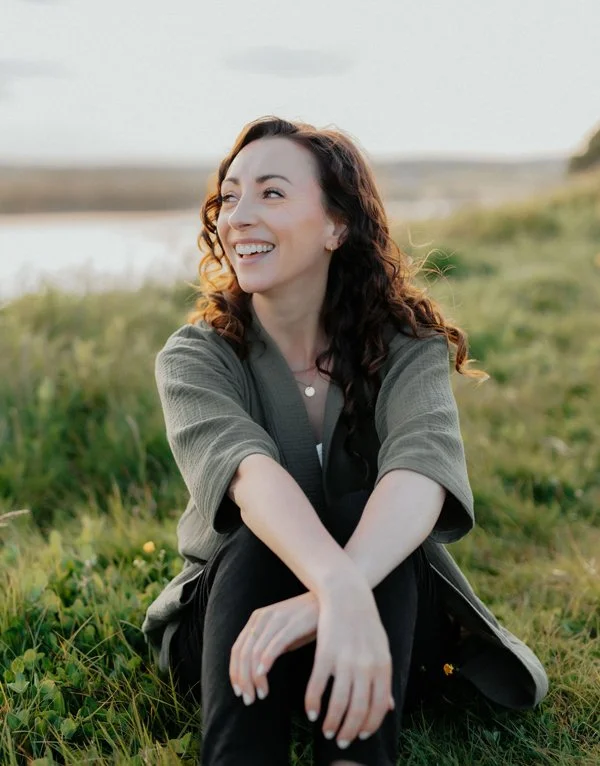 Woman with curly brown hair is smiling and looking off-camera. She wears an olive green sweater and black pants. She is sitting on green grass.