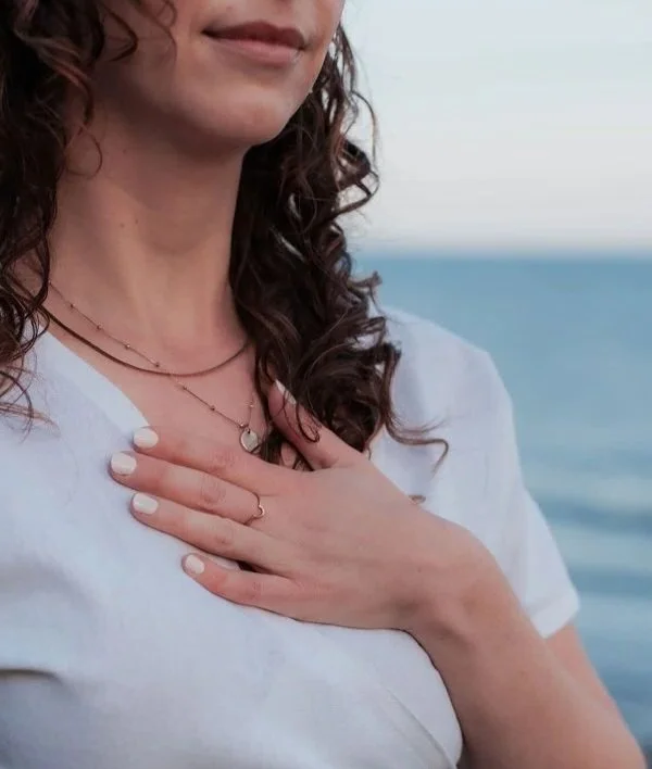 A woman with curly hair wearing a white t-shirt, jewelry, and nail polish, placing her hand on her chest with an ocean background.