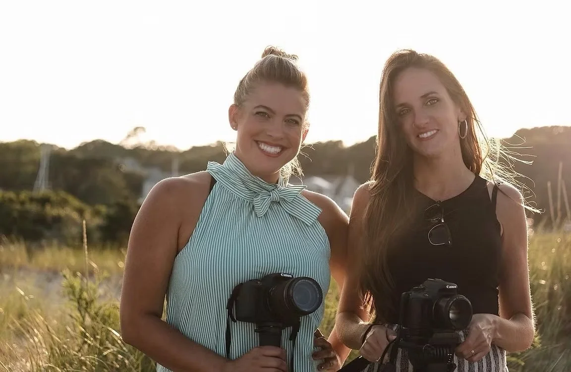 Two women standing outdoors in a grassy area, smiling, each holding a camera, with sunlight glowing behind them.