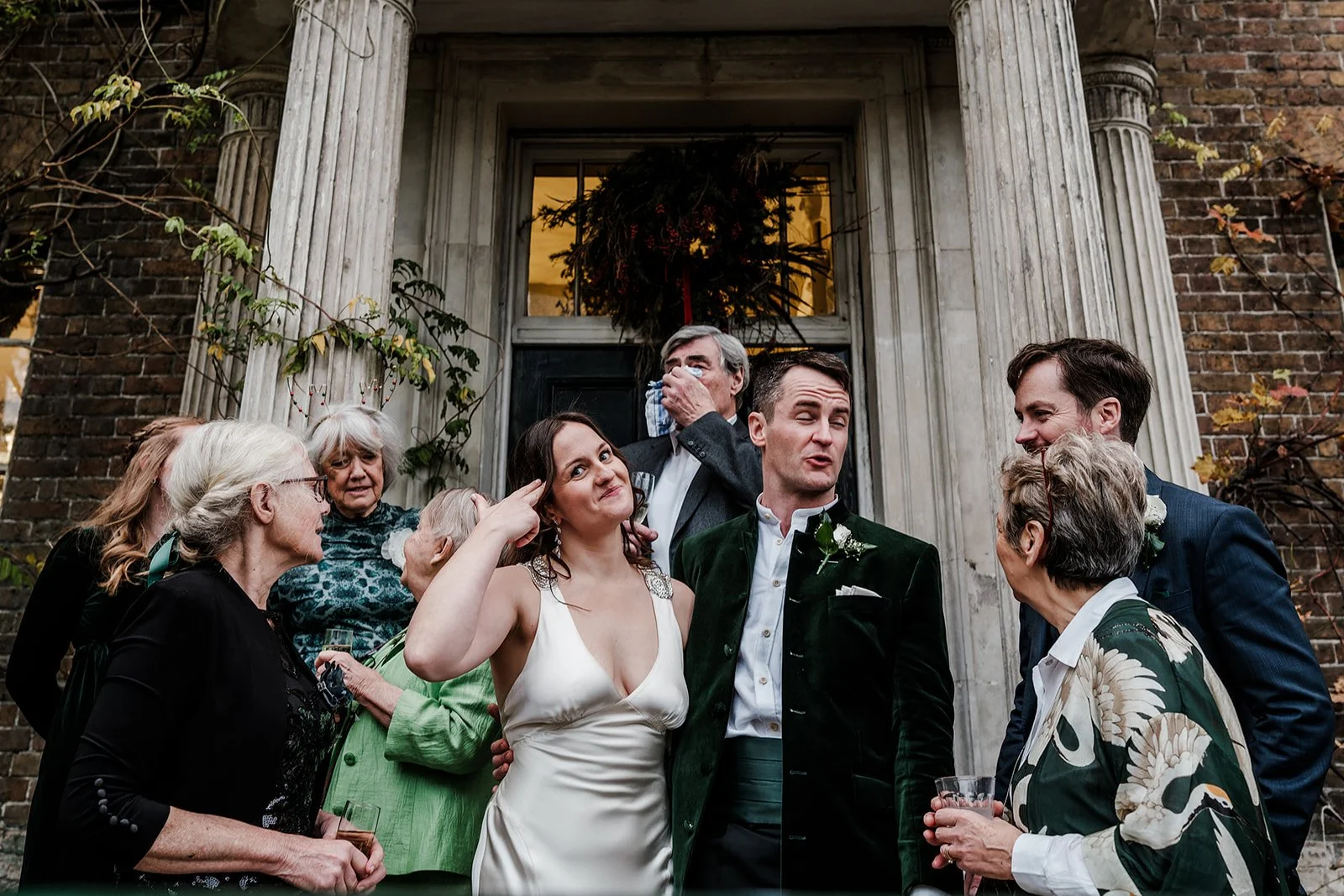A wedding gathering of people standing outside a building with columns, with some guests cuddling and others gesturing, and a couple in the center dressed in wedding attire.