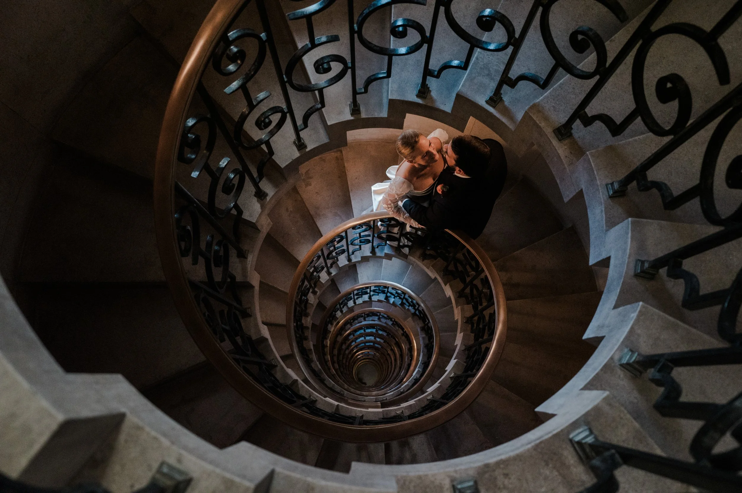 A couple in wedding attire sharing a kiss on a spiral staircase viewed from above.