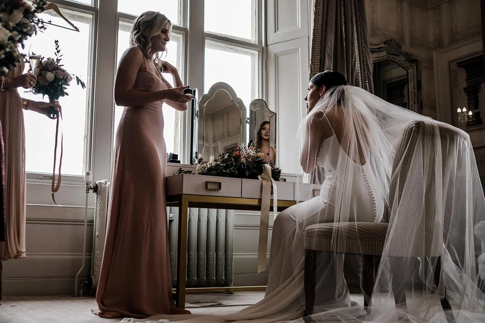 A bridesmaid lifting Sarah&rsquo;s veil by the window at Clevedon Hall, North Somerset
