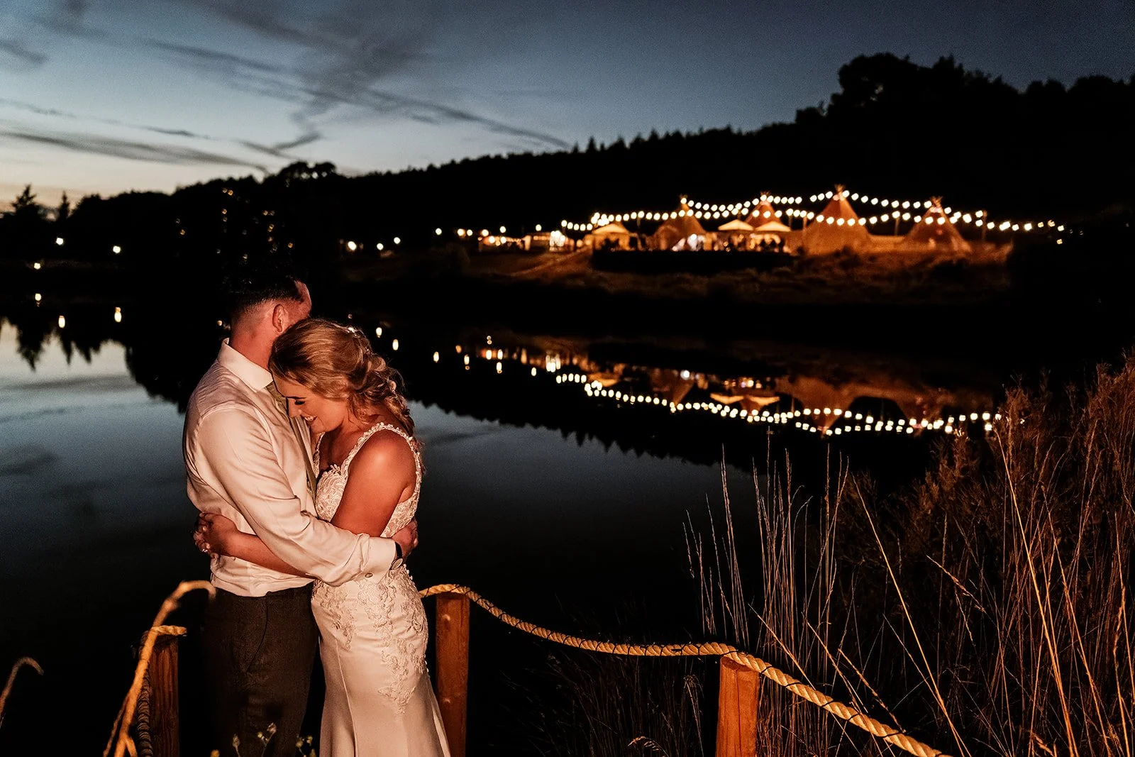 Liv and Jack kissing beside the lake with the tipis lit behind them at Hadsham Farm