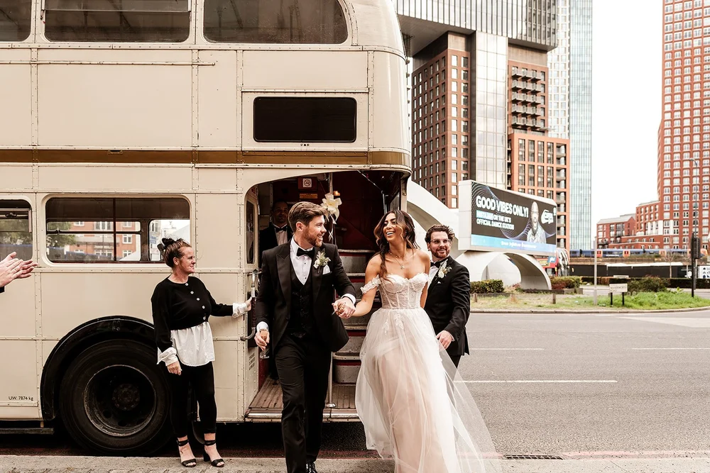 Bride and groom stepping down from a vintage bus by the Thames after their Chelsea wedding