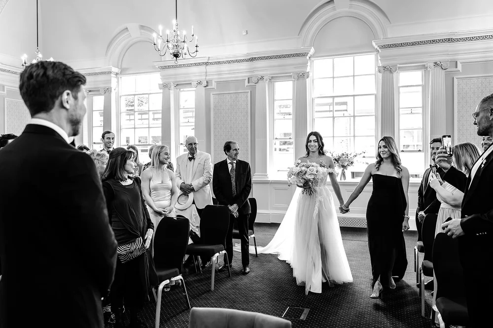 Bride walking into a bright ceremony room with tall arched windows at Chelsea Old Town Hall