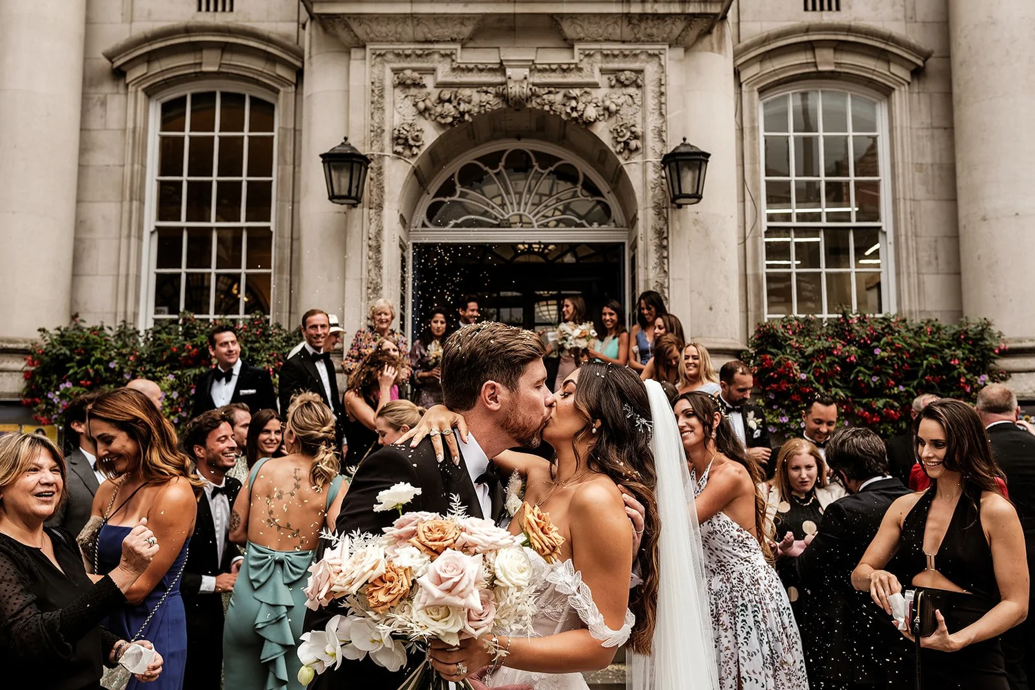 Newly married couple kissing on the steps of Chelsea Old Town Hall surrounded by their guests