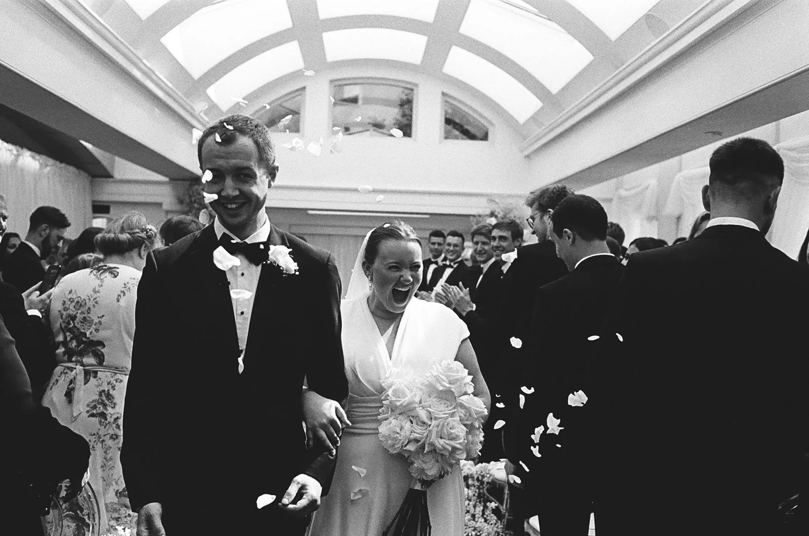 Bride and groom walking through their guests beneath a glazed roof, photographed on 35mm black and white film at a London wedding