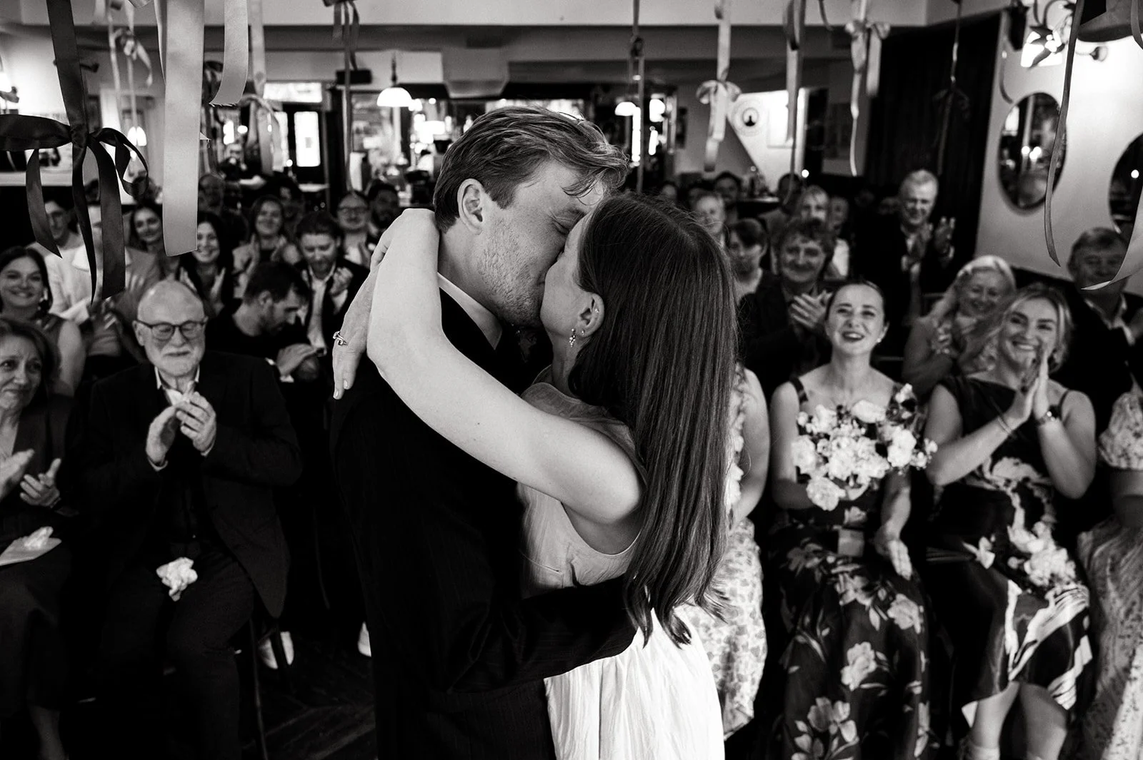 Black and white photograph of Patrick and Ruby kissing surrounded by their guests