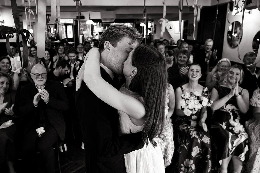 Black and white photograph of Patrick and Ruby kissing surrounded by their guests