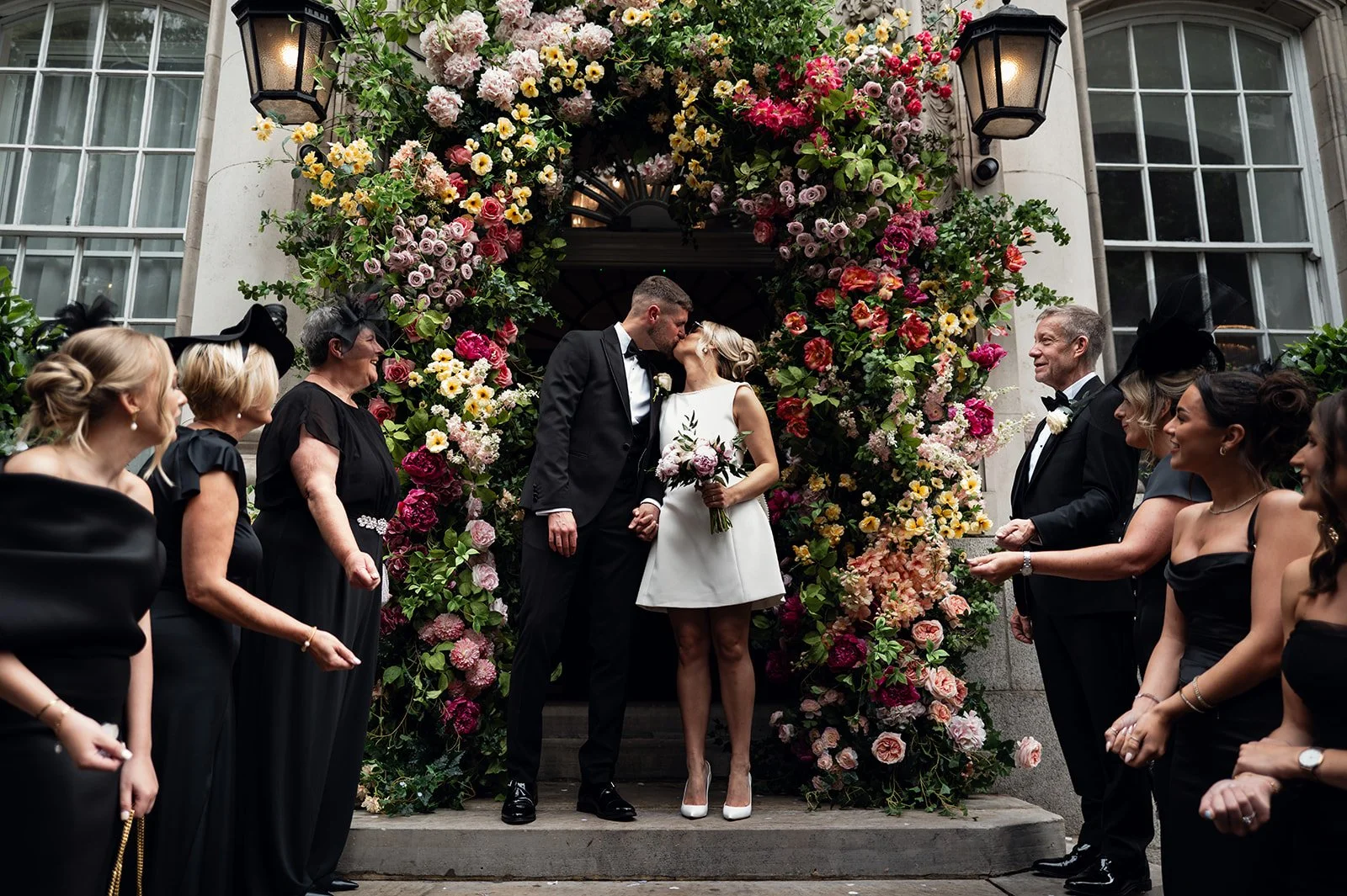 Charley and Joe kissing beneath a flower-covered doorway in Chelsea with their guests either side
