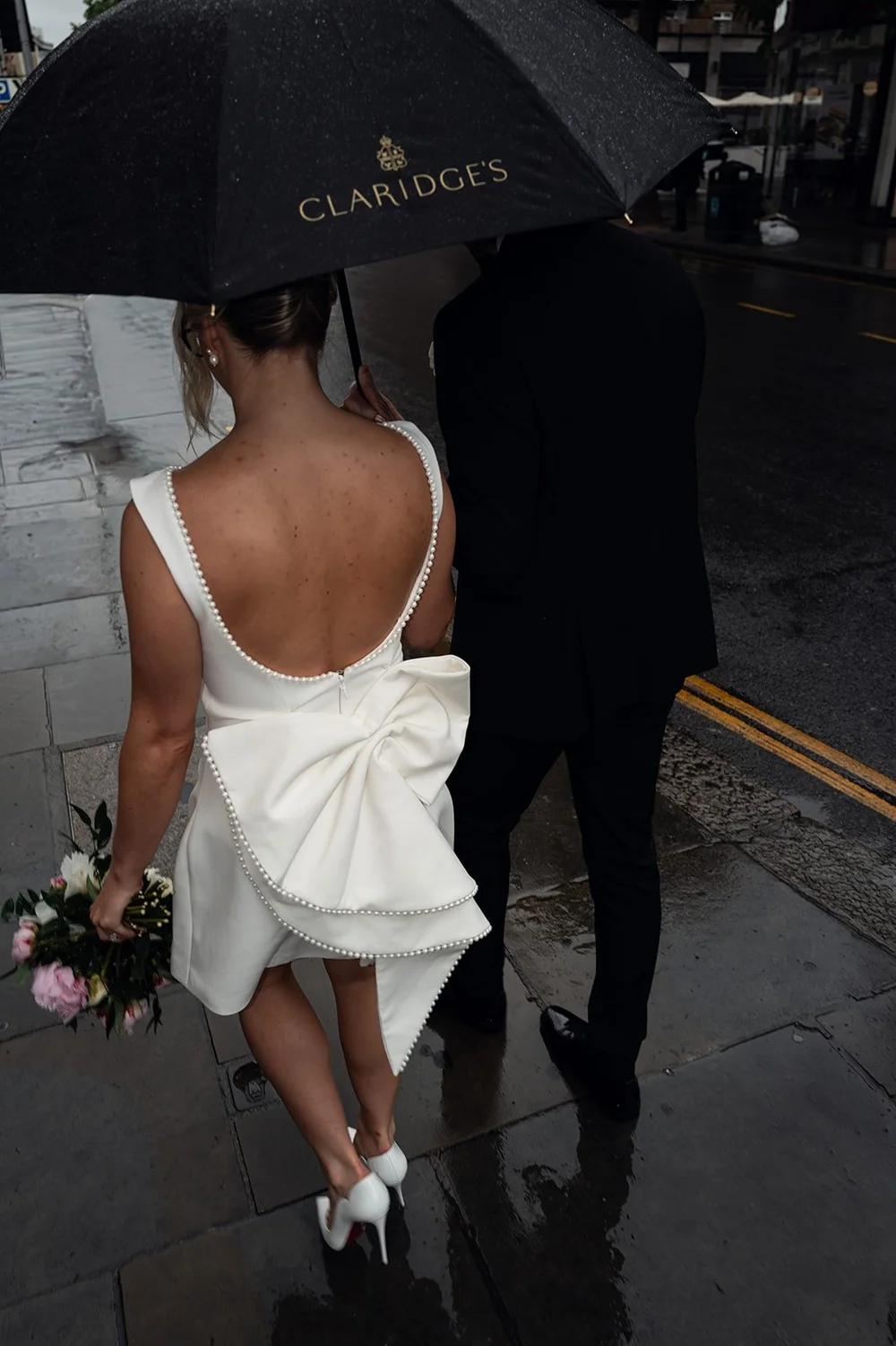 Charley walking through the rain under a Claridge&rsquo;s umbrella in her wedding dress