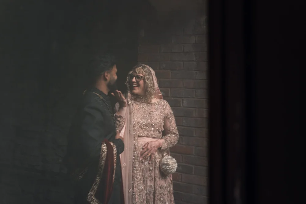 Bride and groom lit softly against a dark wall as they steal a quiet moment