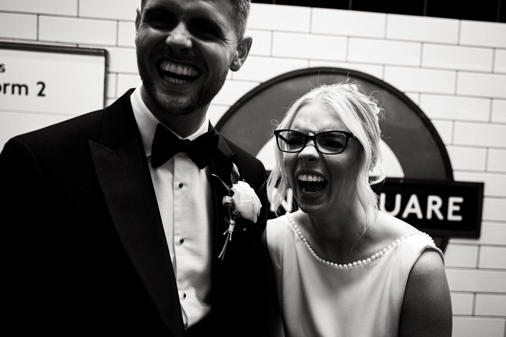 Charley and Joe laughing on the platform at Sloane Square station after their Chelsea Old Town Hall wedding