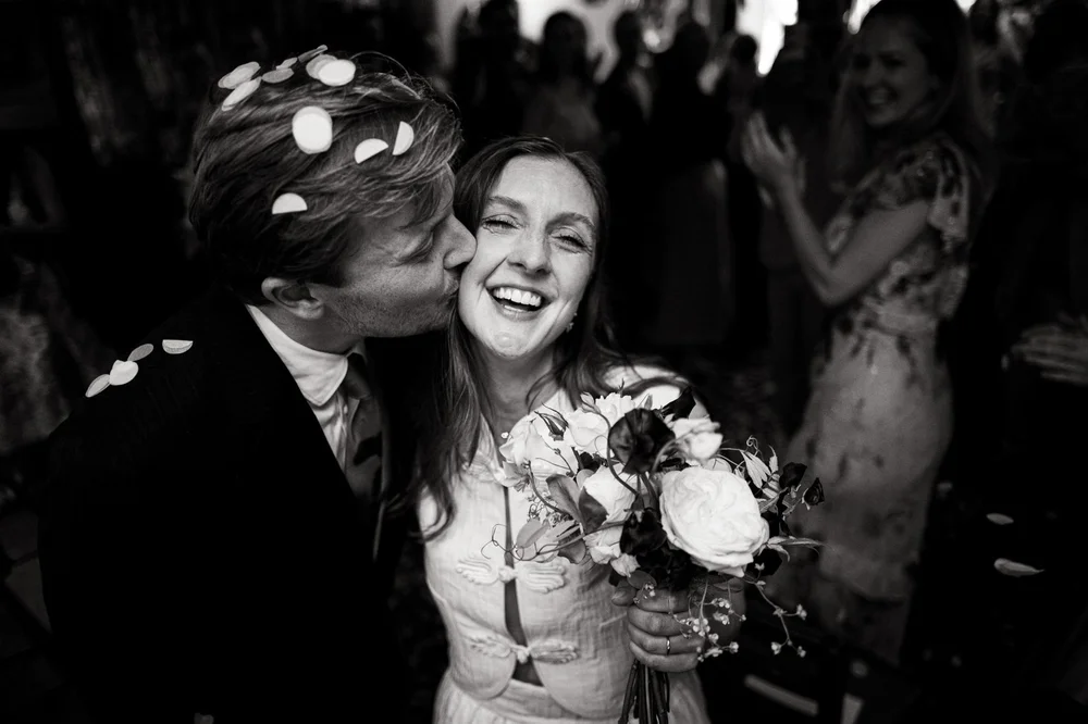 Bride laughing as confetti falls around her while her groom kisses her cheek