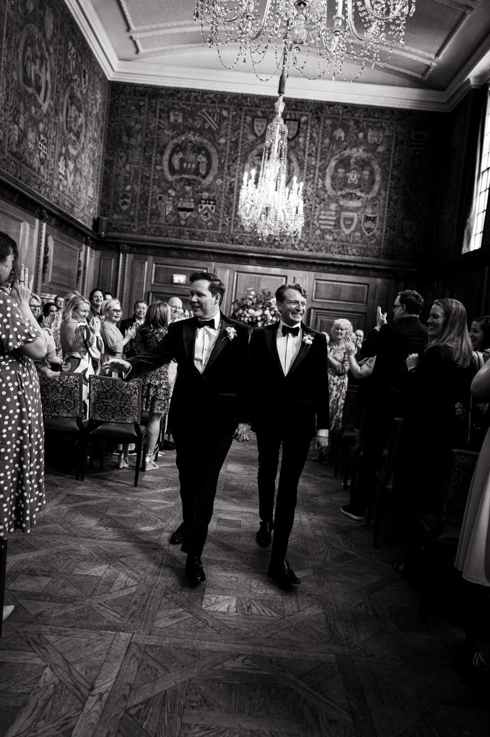 Two grooms walking back down the aisle beneath a chandelier