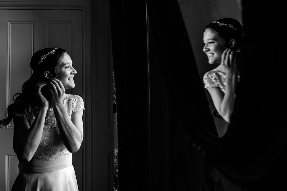 Bride laughing beside a window and reflected in a mirror as she gets ready