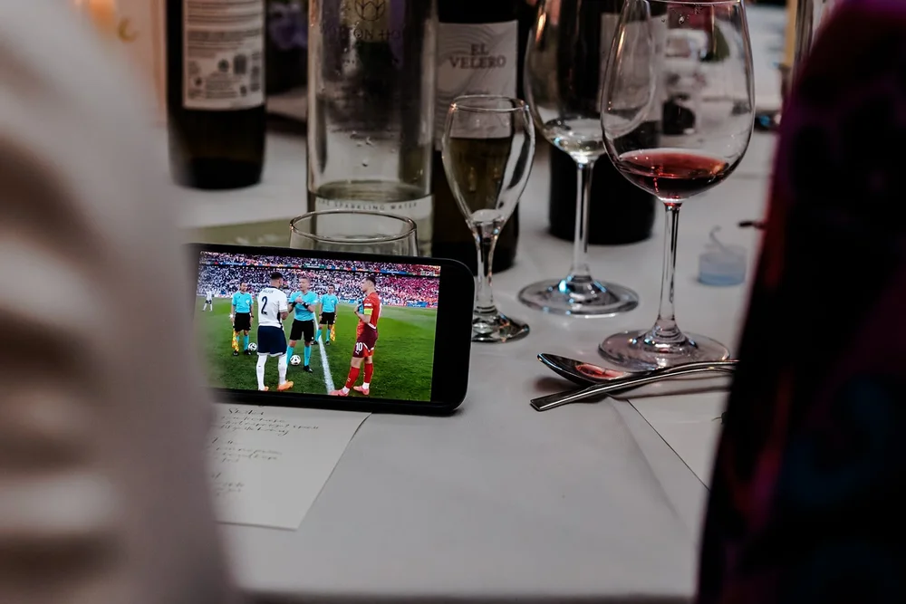 A phone propped on the wedding breakfast table showing a football match between the wine glasses