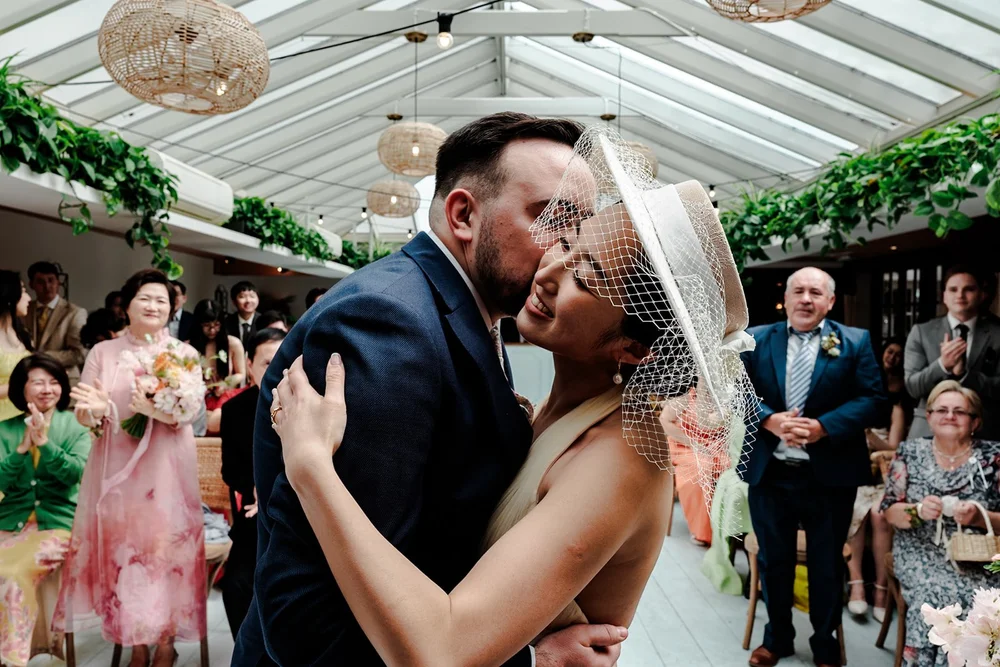 Bride and groom embracing in a glasshouse ceremony room as their guests watch