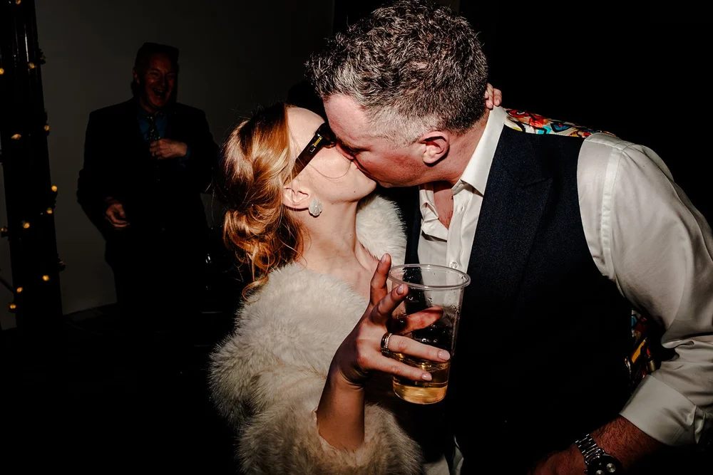 Bride and groom kissing in the middle of the evening party, drinks in hand