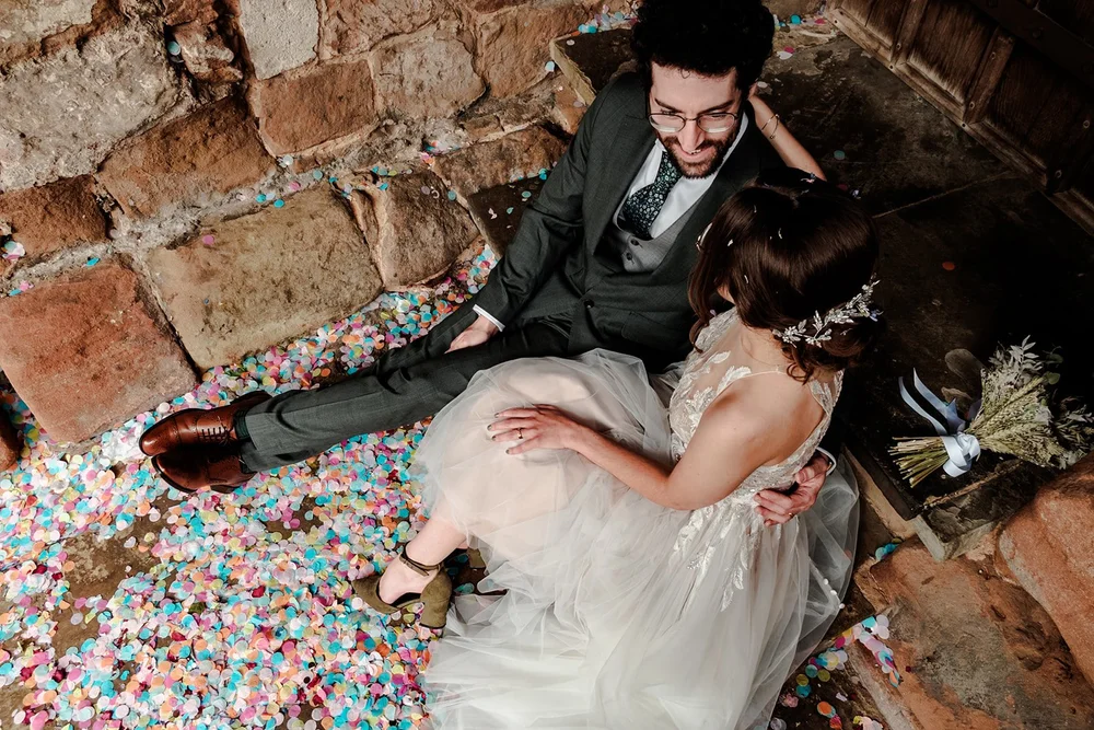 Bride and groom sitting on confetti covered steps against an old brick wall