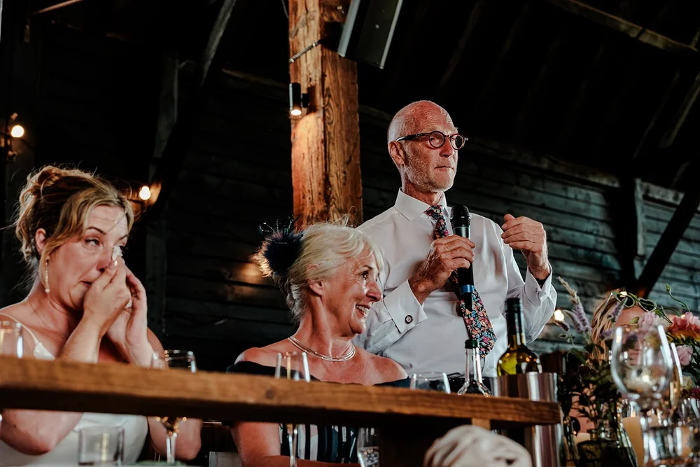 A guest giving a speech in a barn as the bride wipes away a tear