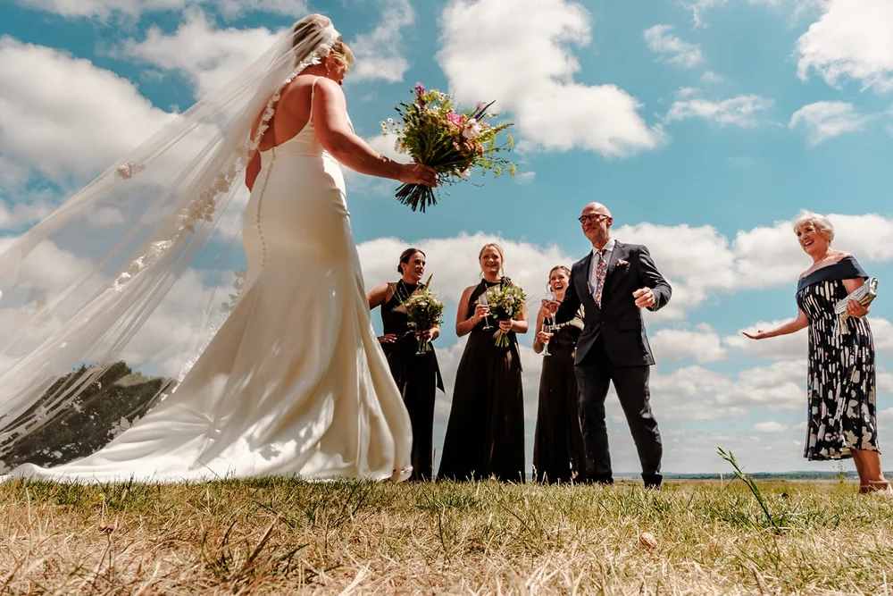 Bride holding her bouquet aloft, veil flying, with her wedding party behind