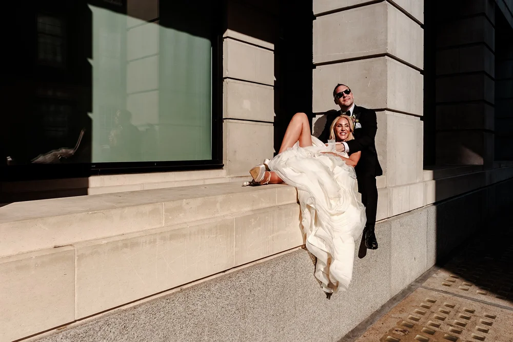 Bride lying back across a stone ledge on a city street beside her groom