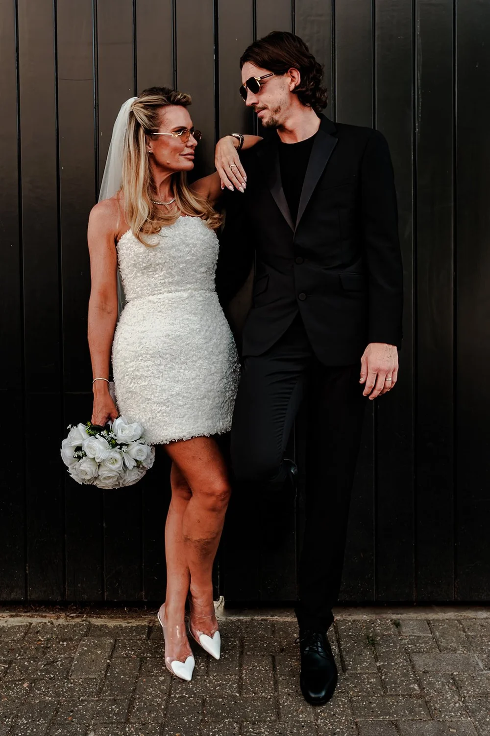 Bride in a short dress and groom in black leaning against a dark wall