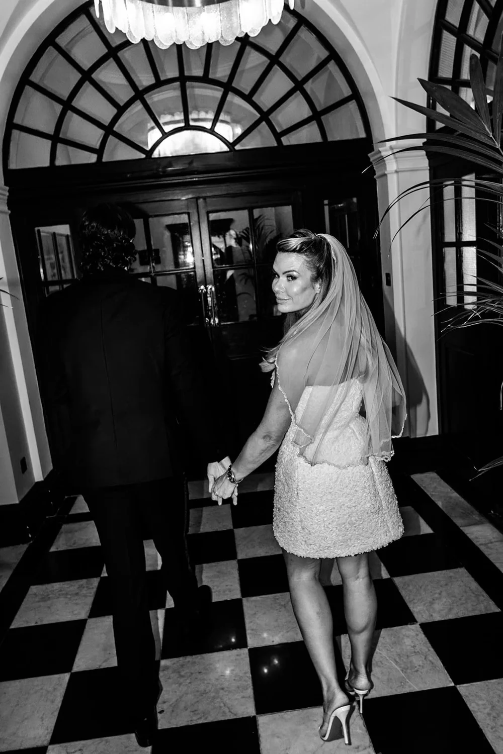 Bride and groom walking across a black and white tiled floor beneath a chandelier