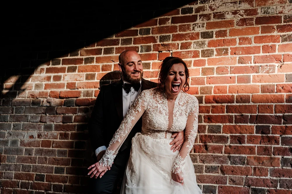 Bride laughing beside a red brick wall with her groom holding her