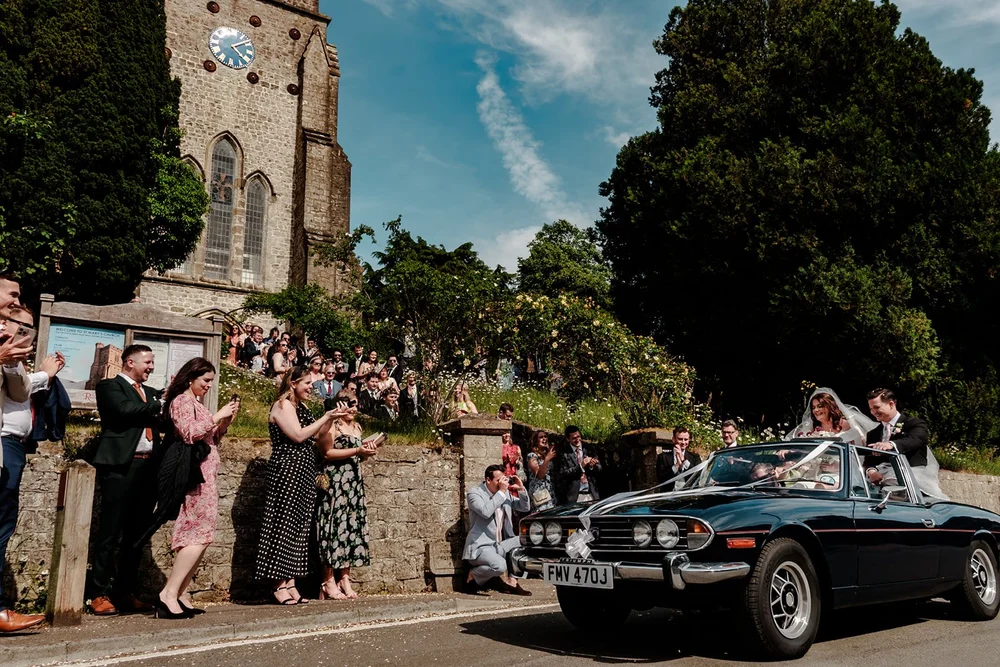 Bride and groom leaving in a classic car as guests line the church wall