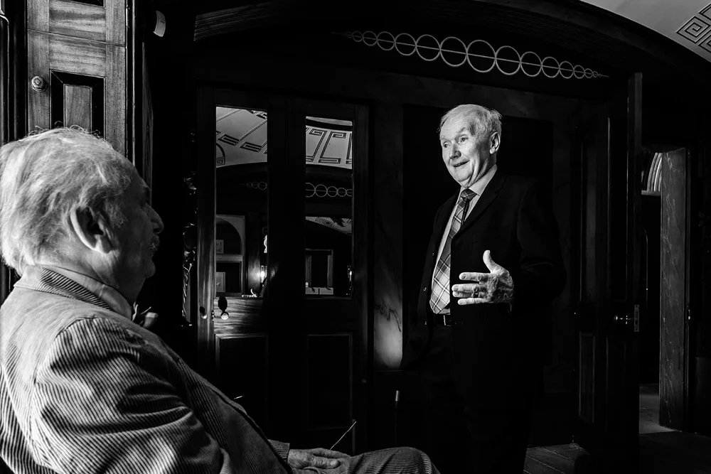 Two older guests deep in conversation in a dark panelled room
