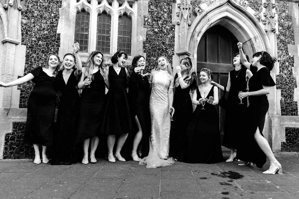 Bride and her bridesmaids in black dresses laughing outside a flint church