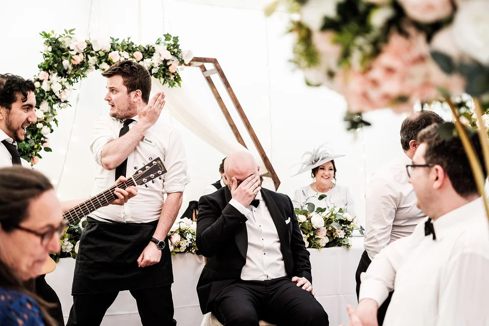 The groom laughing behind his hands as the singing waiters perform