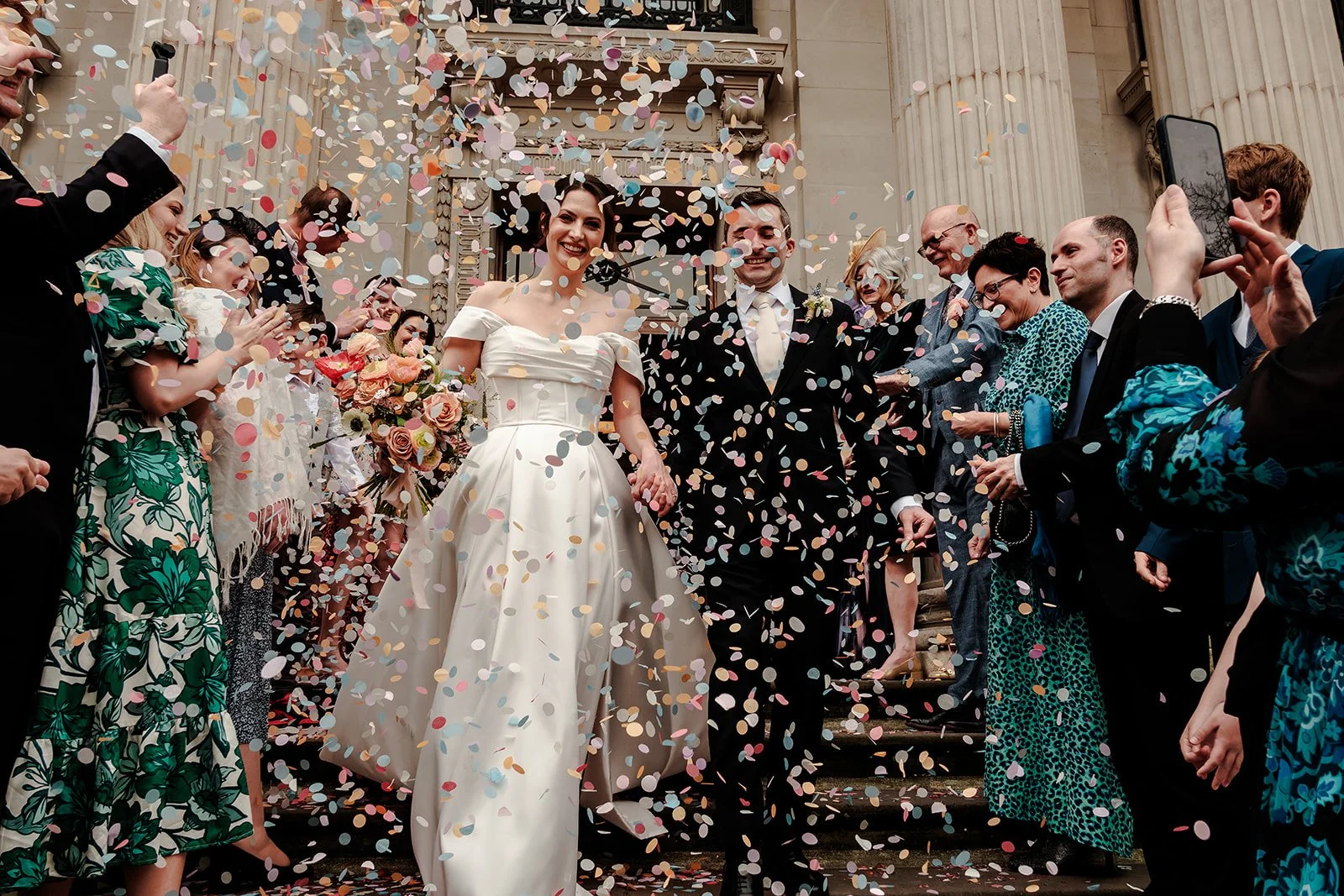 A newly married couple holding hands, surrounded by friends and family, with colorful confetti falling around them outside a grand building with columns.