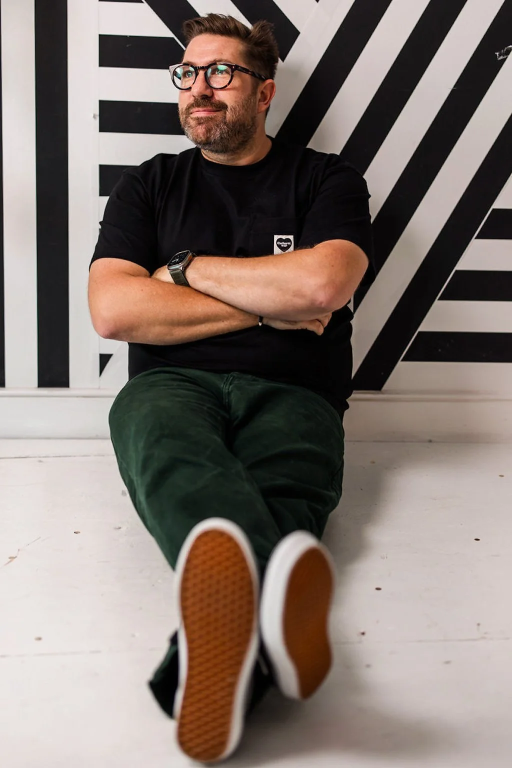 A man with glasses, a beard, and a watch sitting against a wall with black and white geometric patterns, arms crossed, wearing a black t-shirt and green pants, with orange-soled shoes extended towards the camera.