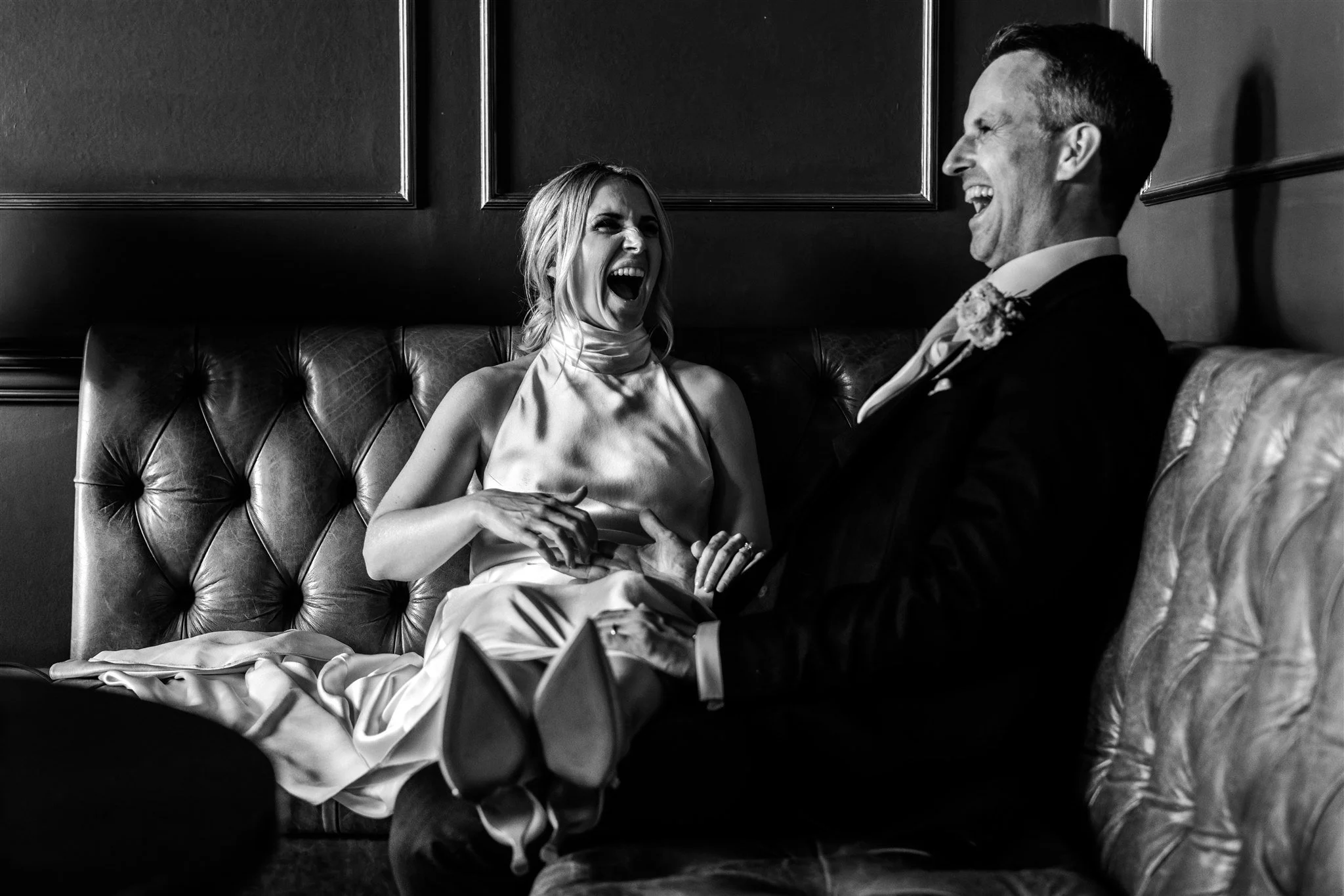 A black and white photo of a woman and man on a vintage leather couch, both laughing and holding hands, woman in a satin dress, man in a tuxedo, in an elegant room.