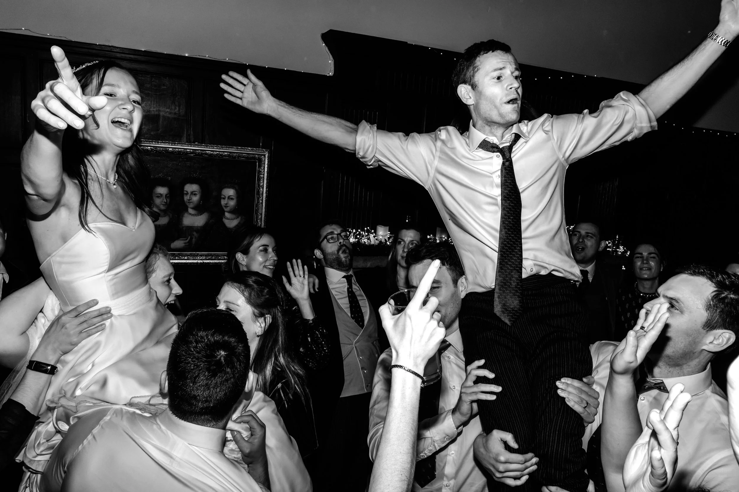 People celebrating at a wedding reception, with a man being carried on someone's shoulders and a woman in a wedding dress dancing happily.