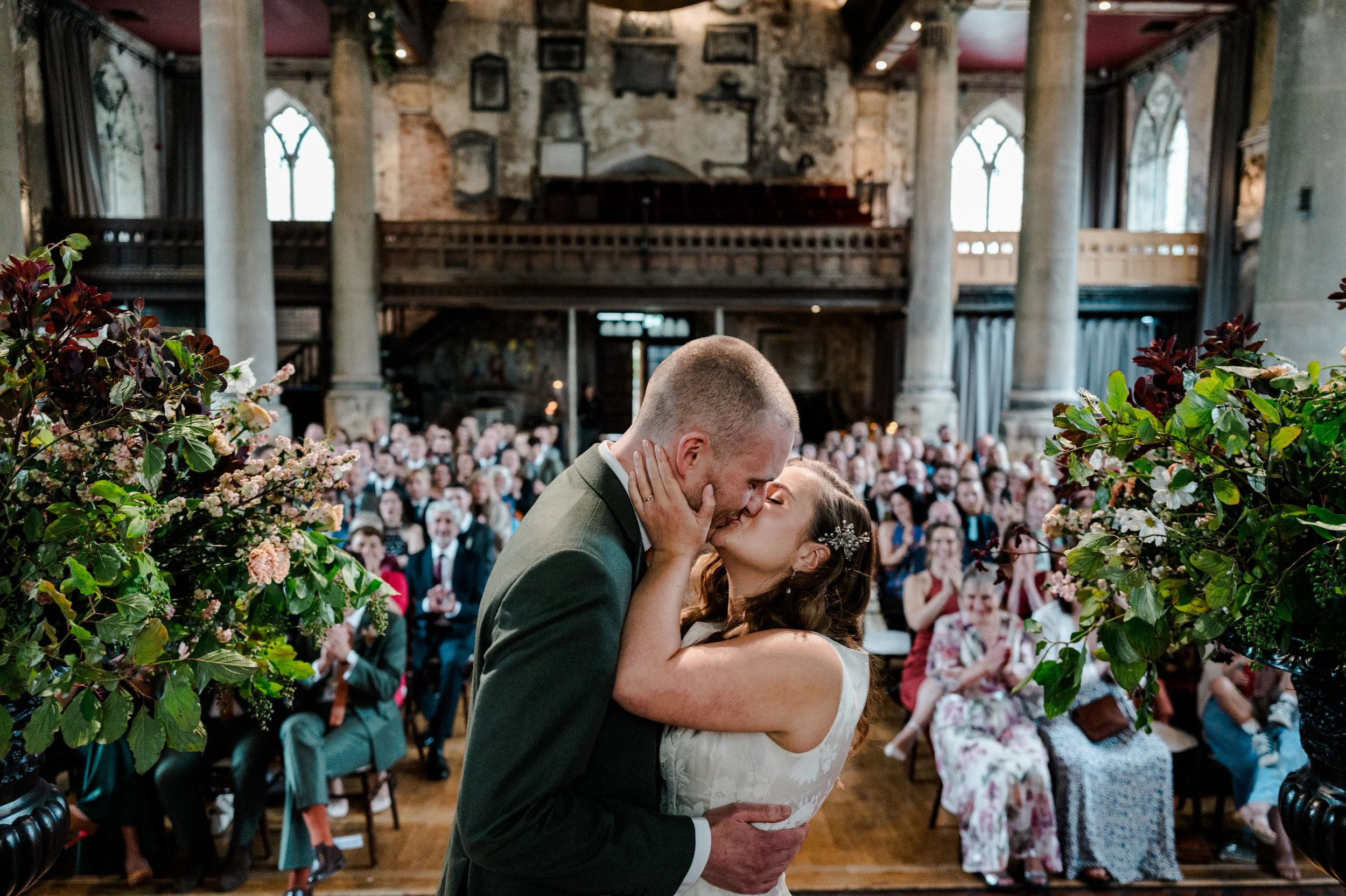 A bride and groom kissing at their wedding ceremony inside a historic church with stone walls, large arched windows, and a crowd of guests applauding in the background. Pink and green floral arrangements decorate the scene.