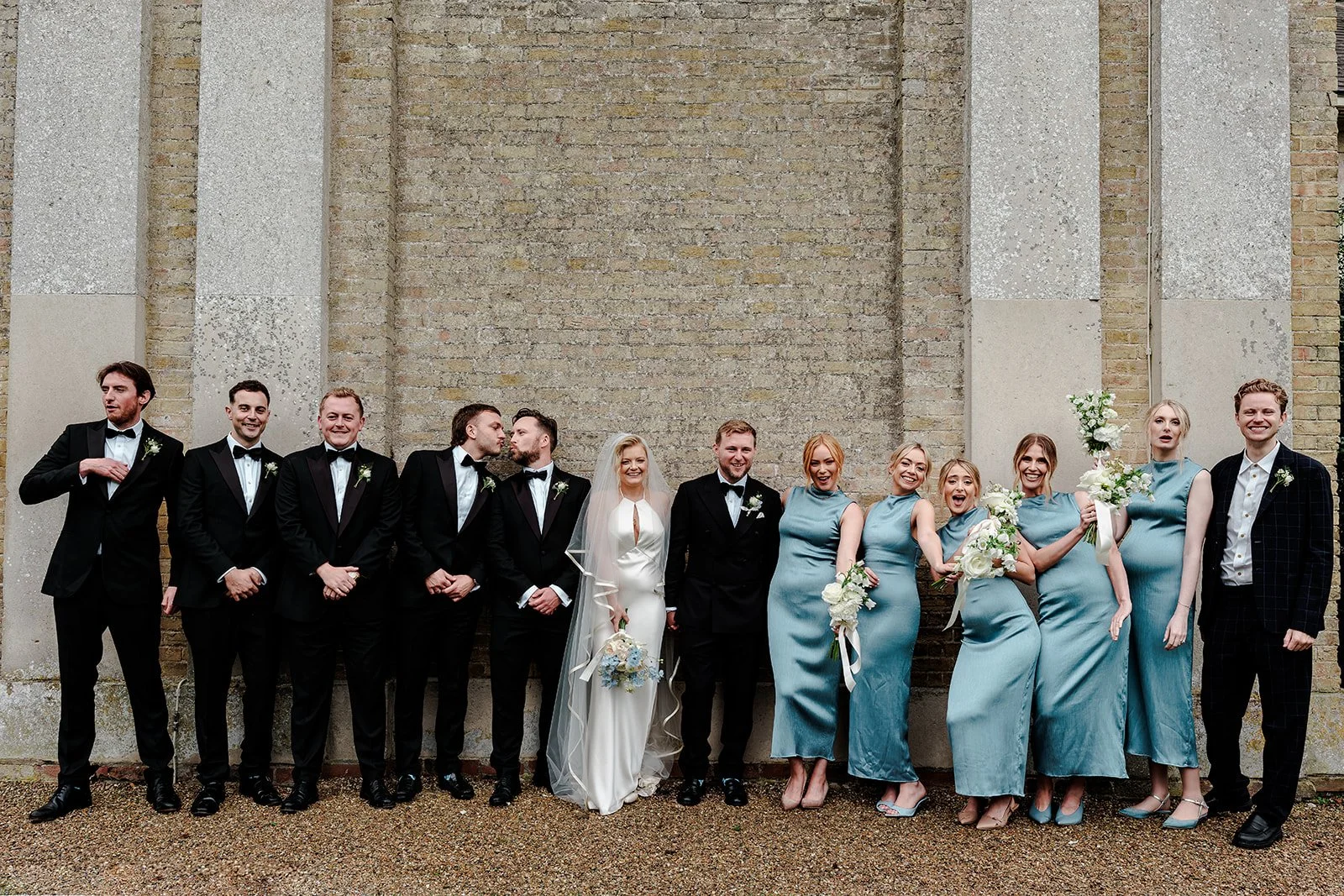 A wedding party with men in black tuxedos and women in blue dresses, standing in front of a brick wall.