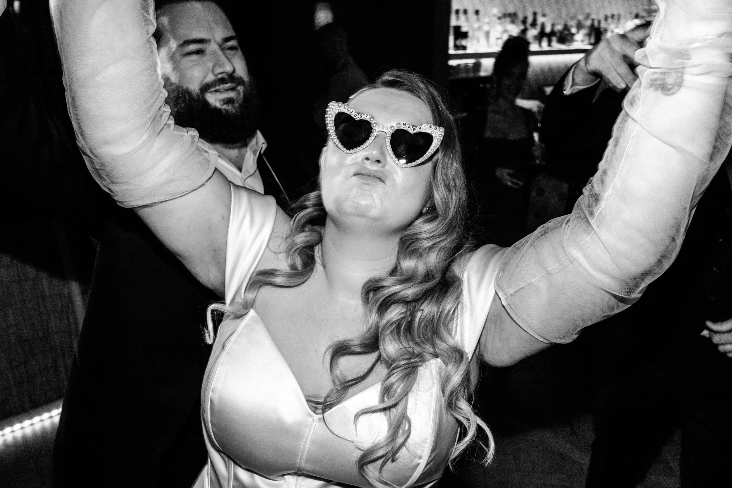 A woman wearing novelty heart-shaped glasses that spell out "just married" and a white dress, dancing with arms raised, at a celebration or party.