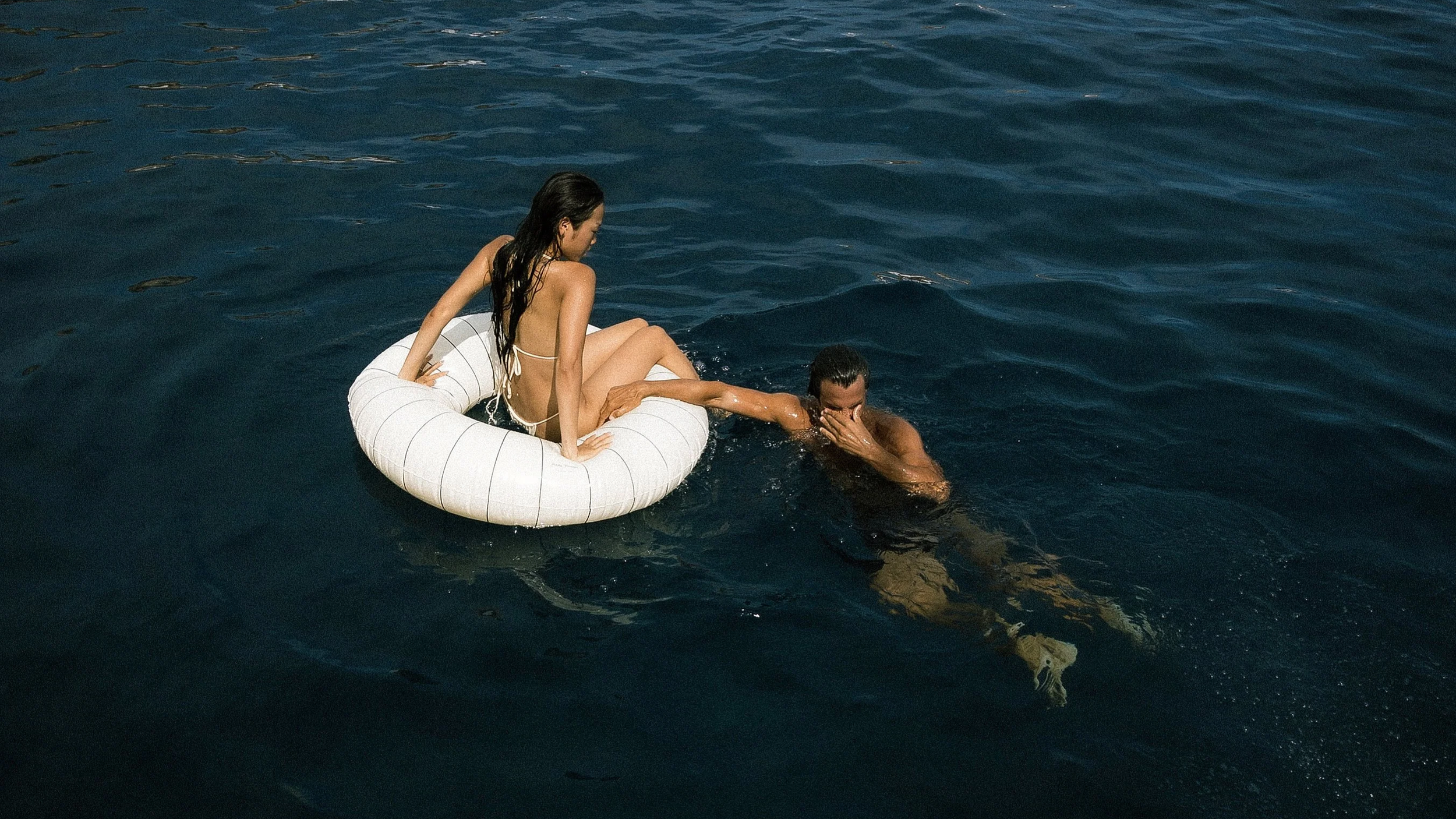 Two women, one sitting on a white inflatable ring and the other swimming in the water, holding hands in a dark blue ocean.