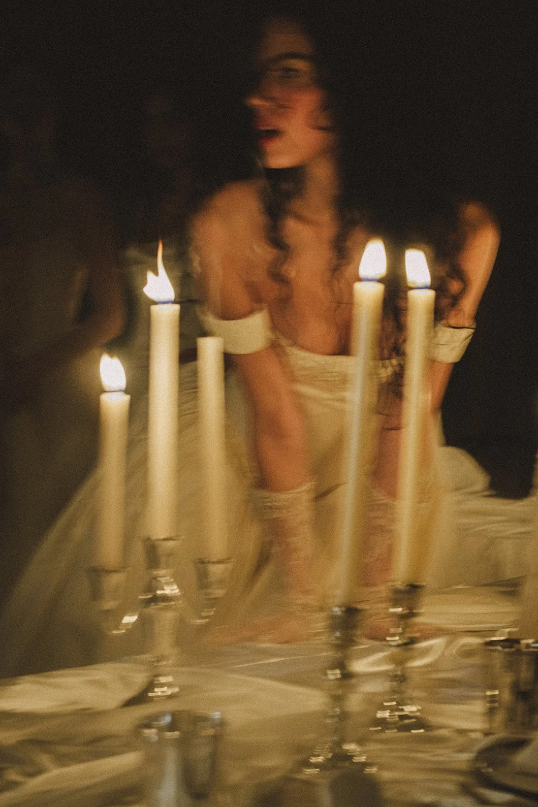 A woman dressed in vintage-style clothing posing behind a candelabrum with lit candles, captured with a motion-blur effect.