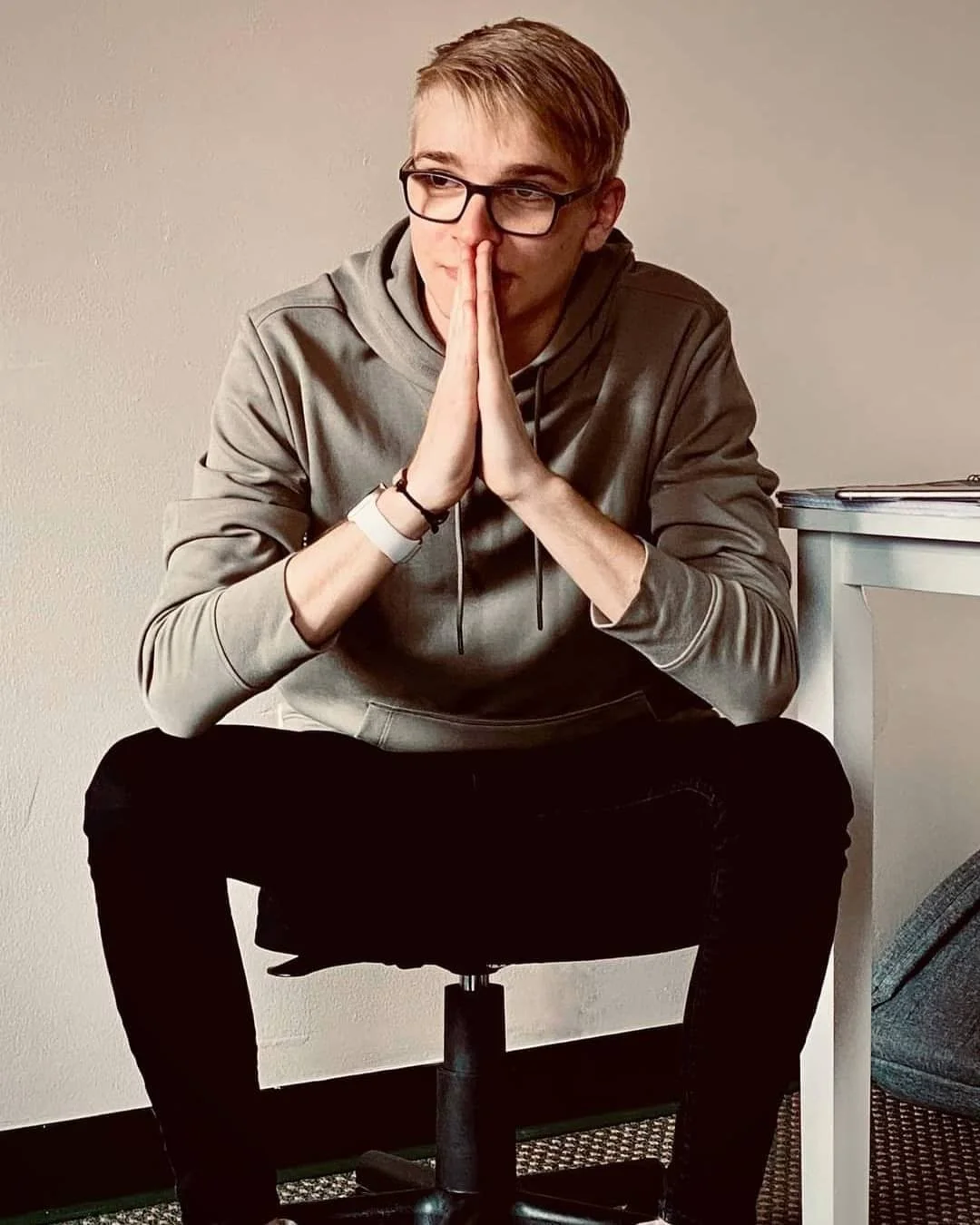 Young man with glasses wearing gray hoodie sitting on a black office chair with hands pressed together in front of his face, sitting next to a gray desk.
