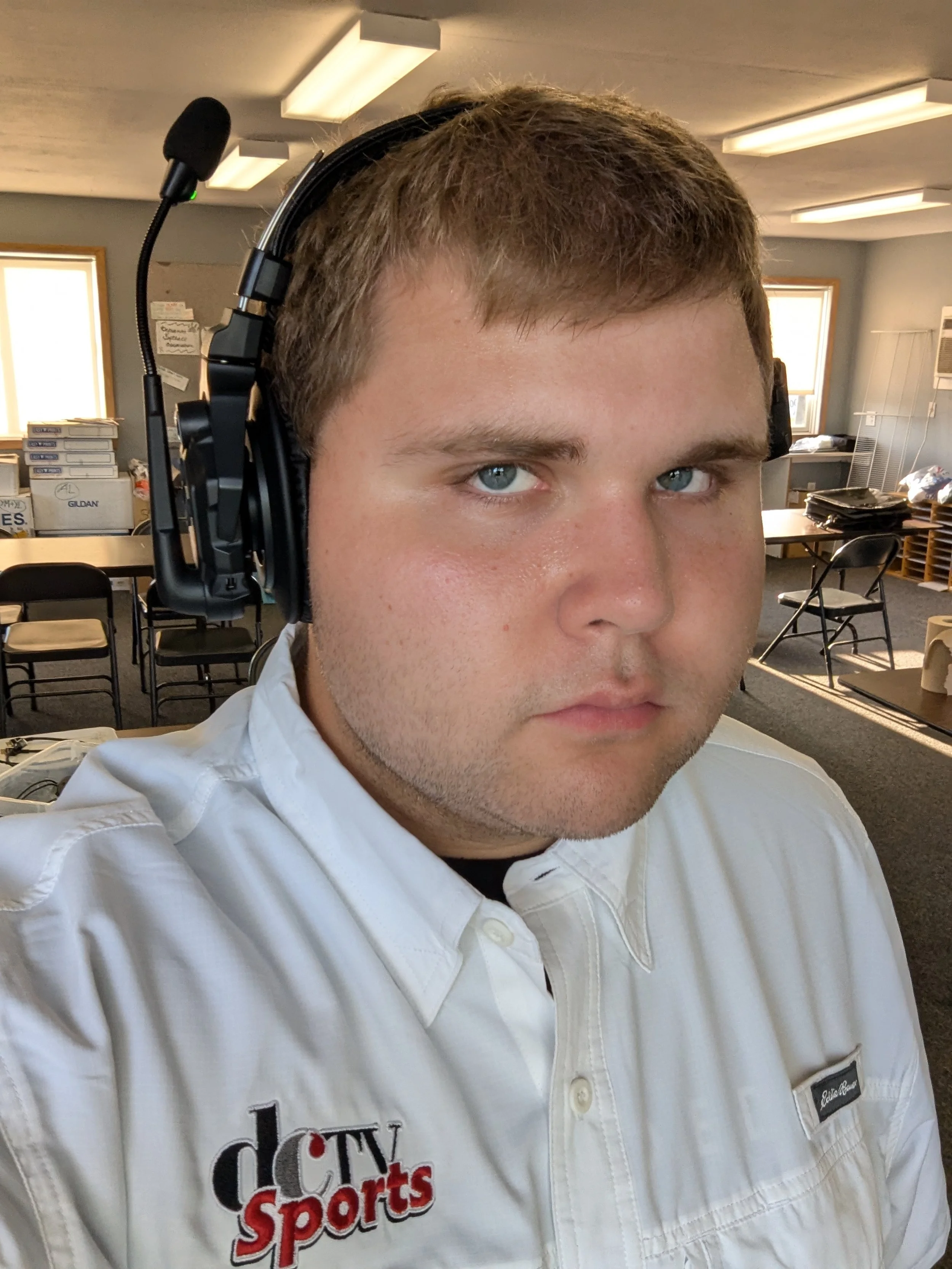 A young man with short brown hair wearing a white shirt with a logo that says 'dCTV Sports' and a black name tag, sitting in a room with tables, chairs, and windows, wearing large over-ear headphones with a microphone.