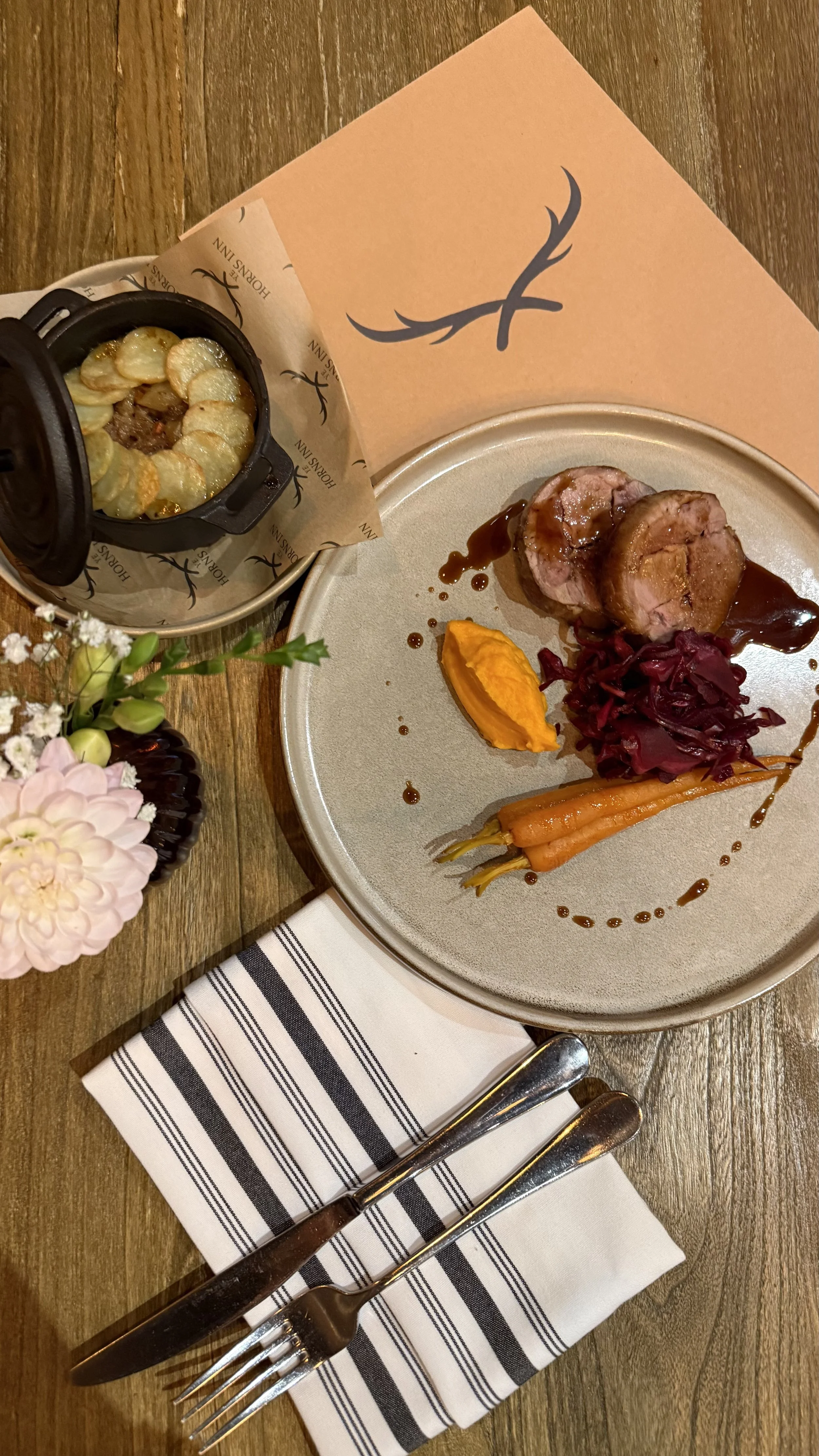 Plate with slices of meatloaf, glazed carrots, mashed sweet potatoes, and red cabbage. Small black ramekin of scalloped potatoes, floral arrangement at side, striped napkin with fork and knife, wooden table.