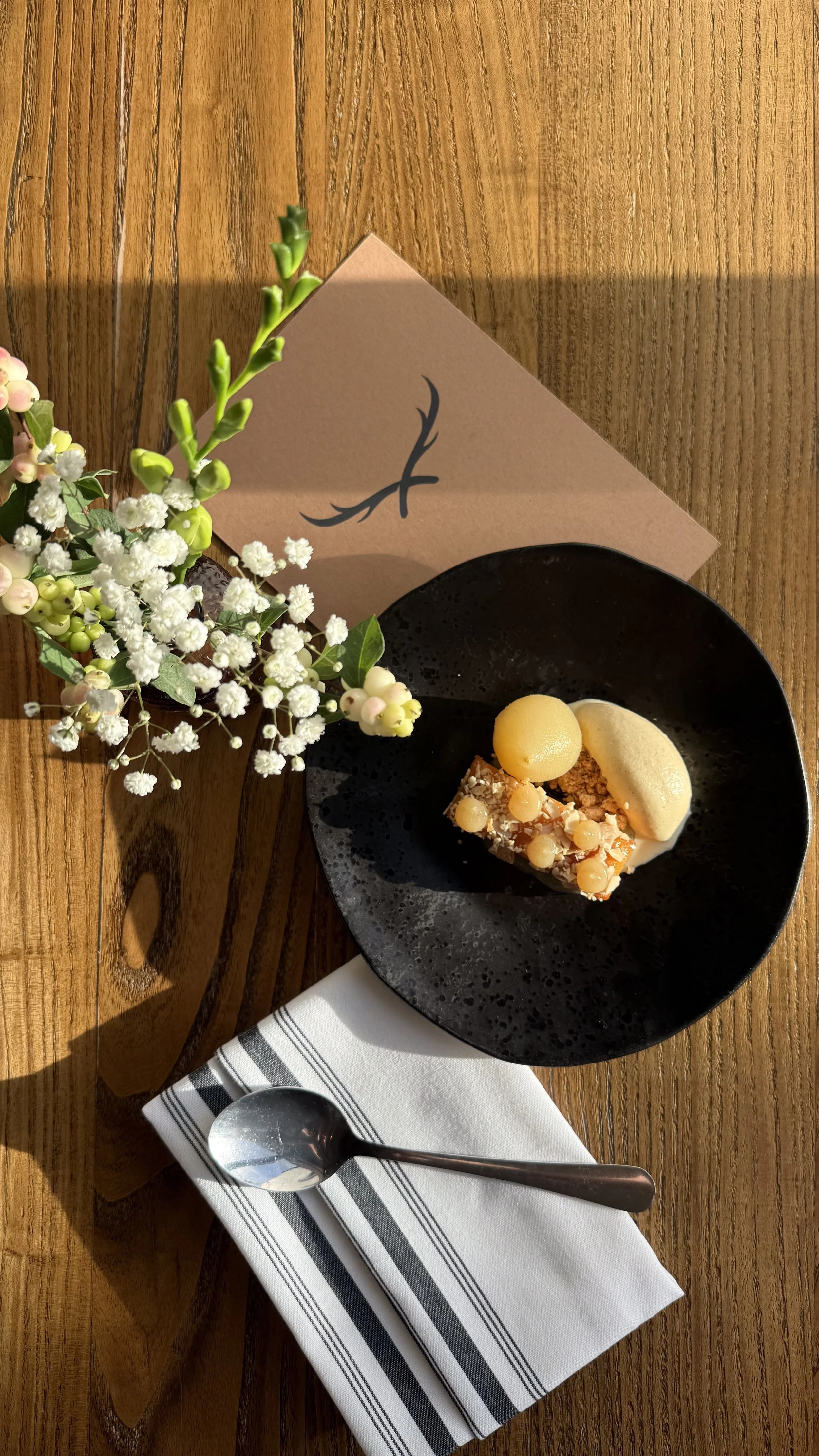 A dessert plate with a slice of cake topped with yellow berries, two round yellow scoops of ice cream, a white scoop of ice cream, and a small white round treat. There is a small vase with flowers and a folded tan napkin with a black design, and a spoon on a striped cloth under the napkin. The background includes a wooden dining table and a pink card with black decoration.