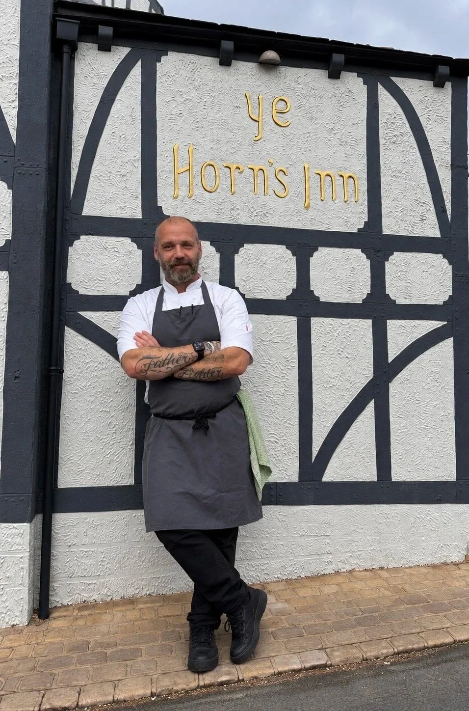 A man with a beard and tattoos, dressed as a chef, stands with arms crossed in front of a building with the sign 'ye Horn's Inn'. He is wearing a white shirt, black apron, and black shoes, with a green cloth hanging from his waistband.