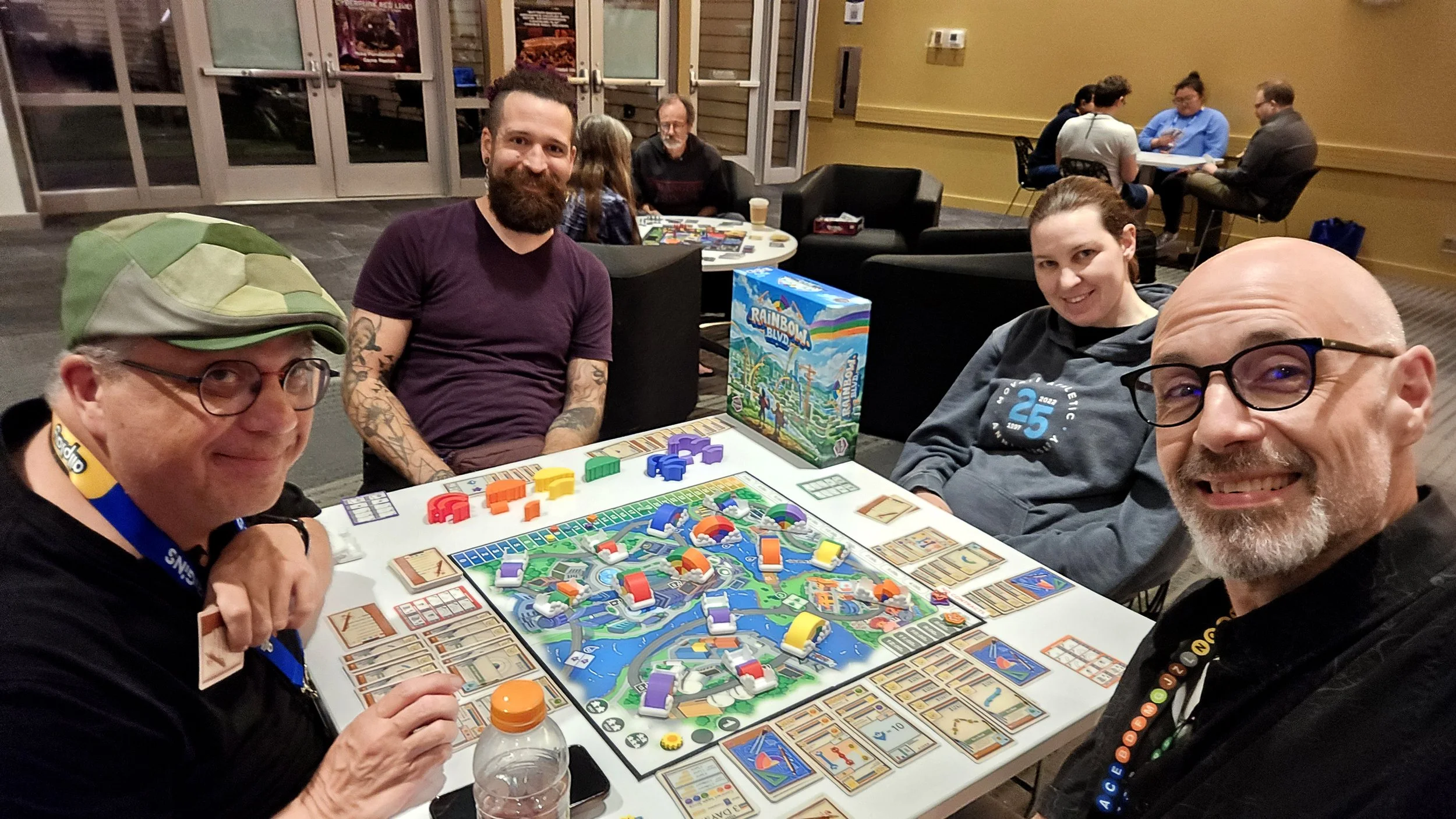 Four people playing Rainbow Blvd, colorful board game at a table, with game pieces, cards, and a game box visible, in a room with other groups in the background.