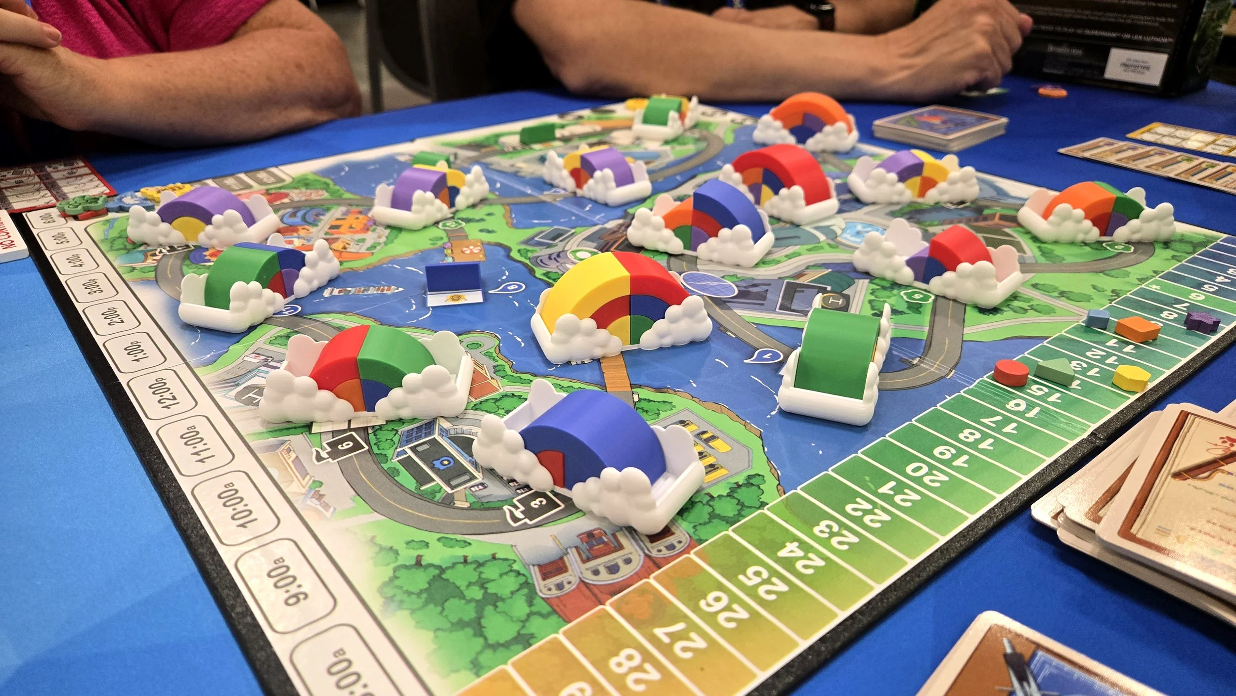 A colorful board game, called Rainbow Blvd., set up on a table with players' arms visible, featuring various game pieces resembling rainbows with clouds, a game board with roads and buildings, and cards included in the game.
